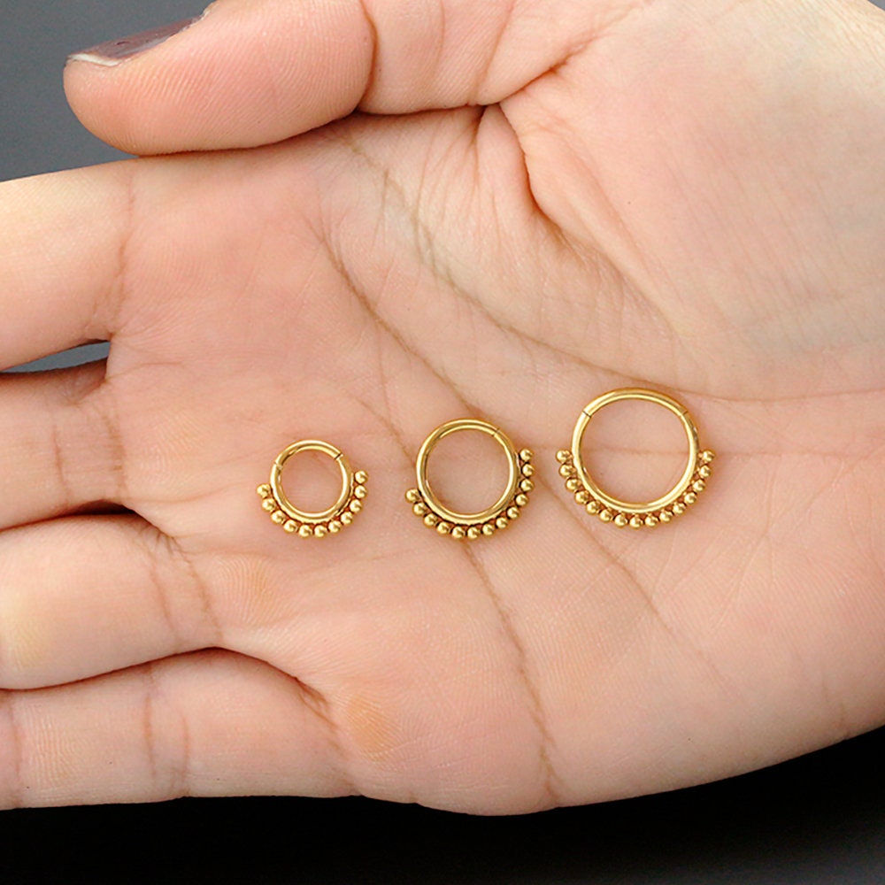 A hand holding three Simple Ethnic Septum rings from Kaya Movement, each crafted in 316L Gold PVD Steel "Etna" and featuring an elegant and traditional design. The rings are neatly arranged in a line from smallest to largest, with small gold beads adorning the bottom edge. The background is a solid dark color, accentuating the intricate ethnic art-inspired design of these 1.2mm gauge, 8mm diameter septum rings.