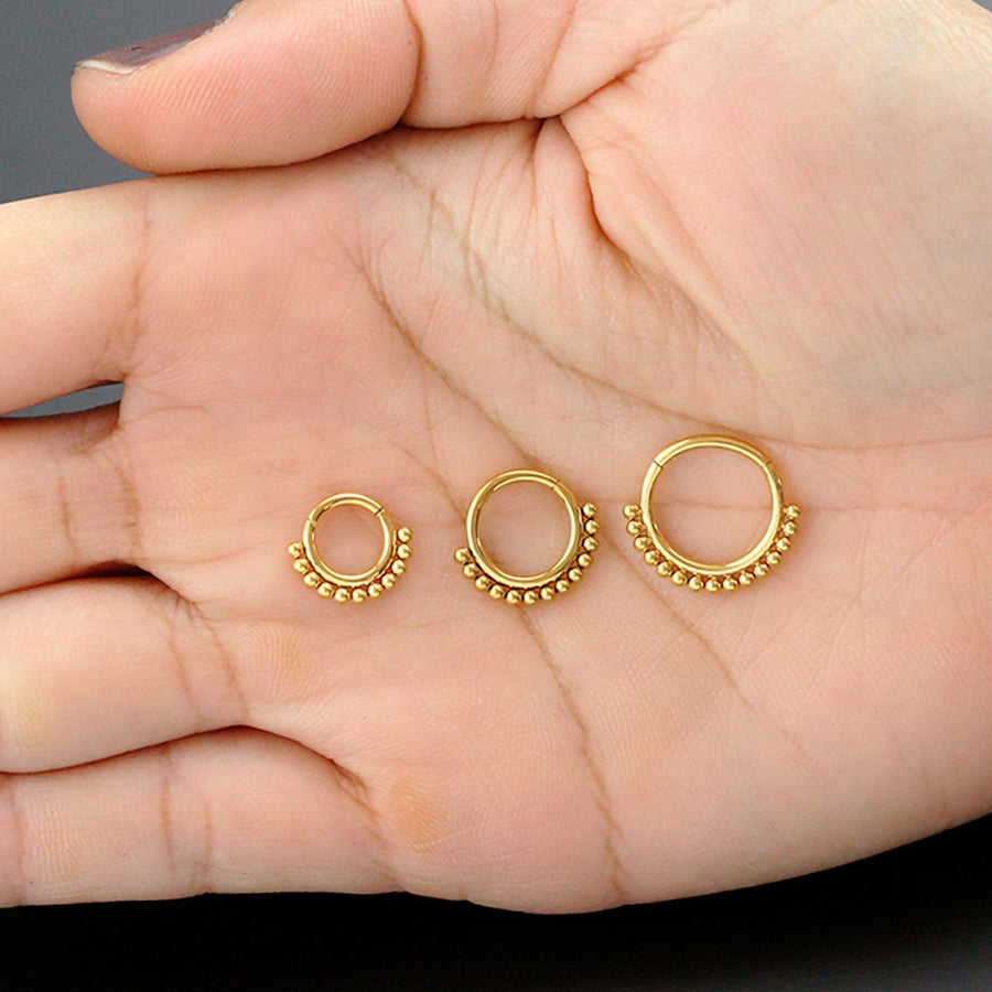 A hand holding three Simple Ethnic Septum rings from Kaya Movement, each crafted in 316L Gold PVD Steel "Etna" and featuring an elegant and traditional design. The rings are neatly arranged in a line from smallest to largest, with small gold beads adorning the bottom edge. The background is a solid dark color, accentuating the intricate ethnic art-inspired design of these 1.2mm gauge, 8mm diameter septum rings.