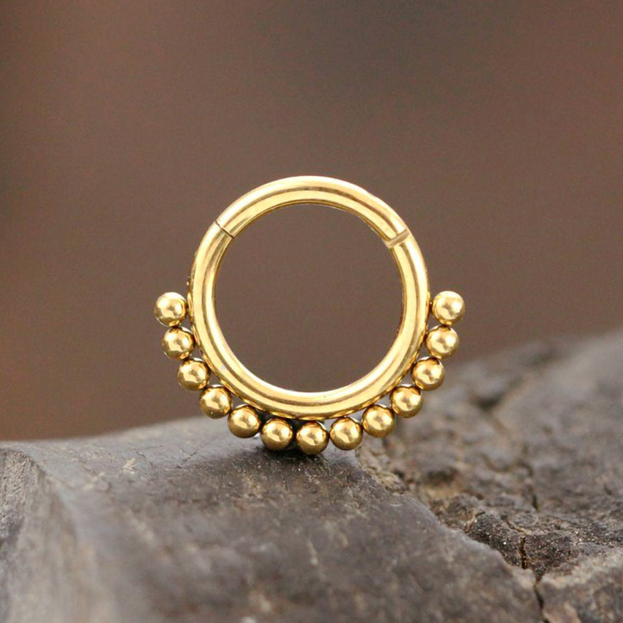 A small, circular gold septum ring from Kaya Movement rests on a textured stone. The lower half of the Simple Ethnic Septum in 316L Gold PVD Steel "Etna," with its elegant and traditional design, is adorned with a series of small, evenly spaced gold beads, adding decorative detail reminiscent of ethnic art. The background is blurred, drawing focus to the intricately designed jewelry piece.
