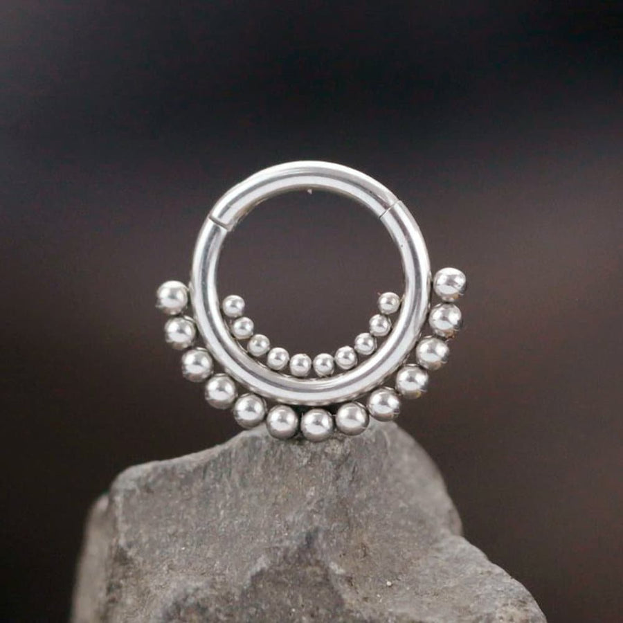 A silver Bohemian Ethnic Septum "Bohra" by Kaya Movement, crafted from 316L steel jewelry and featuring a series of small spherical balls, adorns the lower half. The earring, with a 1.2mm gauge and an 8mm diameter, is positioned upright atop a grey rock against a blurred dark background, accentuating its intricate bead design.