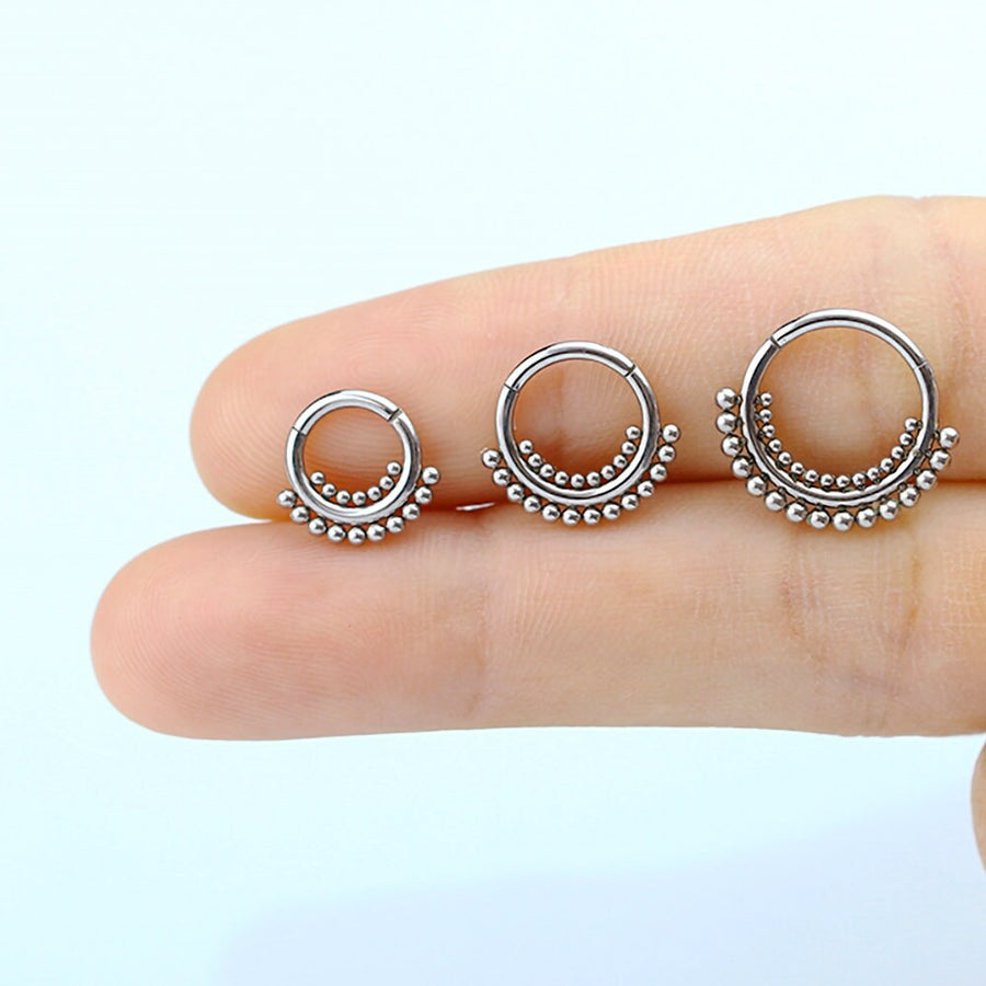 Two fingers hold three Bohemian Ethnic Septum "Bohra" earrings made from 316L steel, each featuring a semi-circle arrangement of small metallic balls along the bottom half. These elegant pieces by Kaya Movement offer a touch of bohemian flair against the plain white background.
