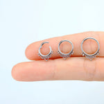 A close-up image of a hand holding three small, circular septum rings from Kaya Movement's "Zura" collection, featuring mandala-inspired designs. The minimalist rings, crafted from 316L steel with a silver look, showcase tiny decorative elements including heart-shaped details at the bottom of each ring. The background is a plain, light blue.