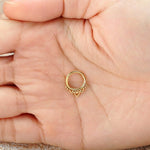 A hand holds a small, intricate piece of jewelry from Kaya Movement. The Minimalist Mandala-Inspired Septum in 316L Steel PVD Gold "Zura" features a heart-shaped design with delicate decorative elements surrounding it. The background is slightly blurred, emphasizing the details of the septum ring and the person's hand.