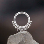 An Aria Minimalist Mandala Septum nose ring in 316L Steel with a silver finish, featuring a 1.2mm gauge and an 8mm diameter, ideal for tragus, lobe, and conch piercings by Kaya Movement, is displayed against a dark blurred background while rested on a rough grey rock.
