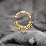 A close-up photo of the Liora Minimalist Mandala-Inspired Septum in 316L Steel Gold Finish by Kaya Movement placed against a blurred, rocky background. The septum jewelry has a circular shape and features small decorative elements, including heart-shaped designs and tiny spherical ornaments along its lower edge.