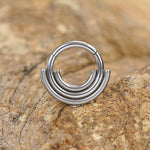 A close-up of the Lira Minimalist Boho Chic Septum by Kaya Movement, featuring a sleek geometric design with three layered semi-circular arcs. Crafted from 316L steel with a refined silver finish, this 1.2mm gauge, 8mm diameter piece embodies modern-day elegance as it rests on a textured beige and brown stone surface.