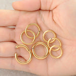 A hand holds several small Aurel Simple Ear Rings from Kaya Movement, each in a 316L Steel Gold PVD Finish. The minimalist hoops, available in gauges of 2 or 3mm and diameters of 8, 10, 12, and 14mm, are arranged in a slightly scattered manner on a neutral light-colored surface. The shiny, evenly-shaped earrings overlap partially, showcasing their elegant simplicity.