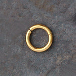An Aurel Simple Ear Ring from Kaya Movement, crafted in 316L steel with a gleaming Gold PVD finish, is displayed on a dark, textured surface. The small, circular hoop earring exudes minimalist elegance and features a smooth, shiny surface along with a subtle hinge mechanism visible on one side. This particular ring is available in gauges of 2 or 3mm and comes in diameters of 8, 10, 12, and 14mm.
