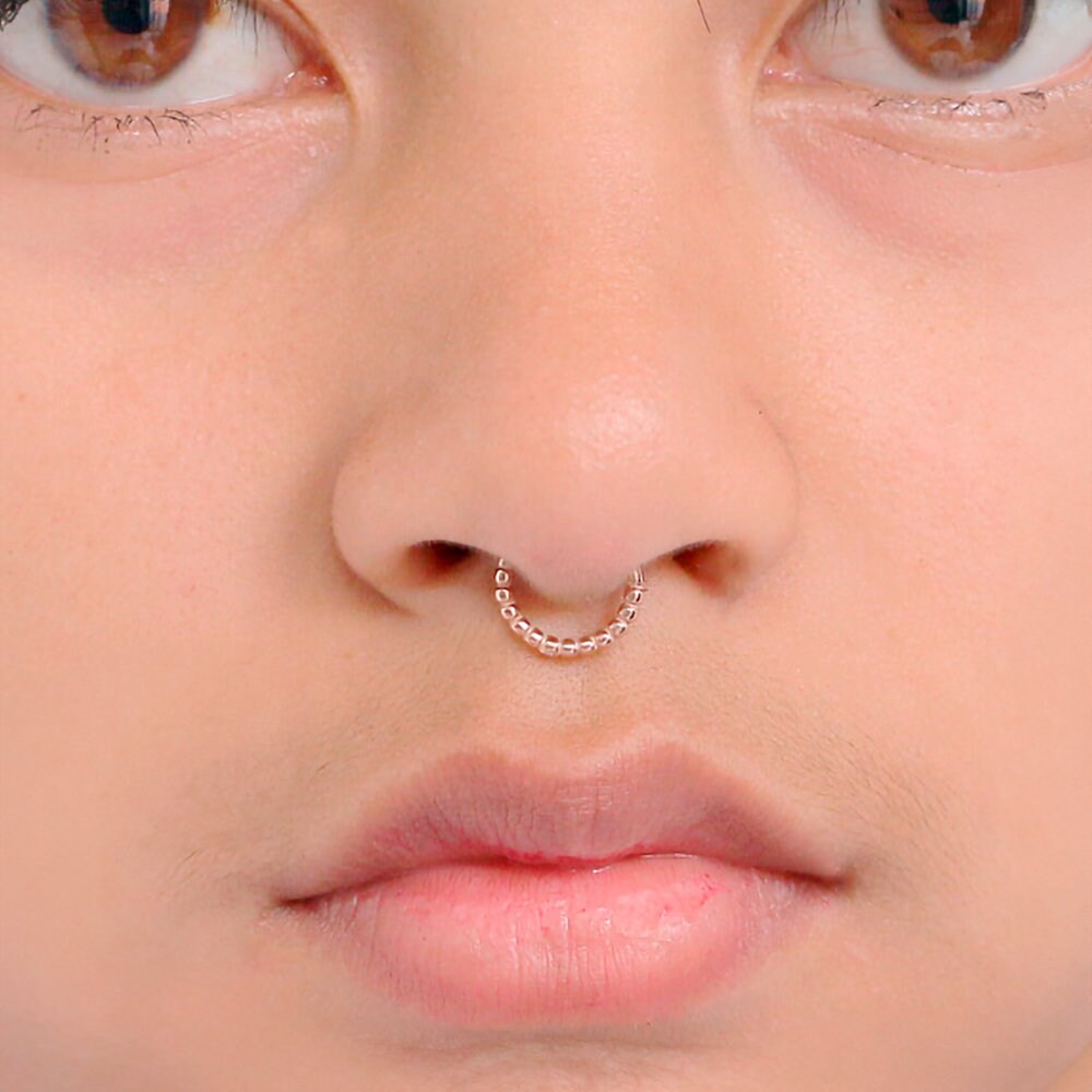 Close-up of a person with light skin wearing the Liora Elegant Septum from Kaya Movement. The delicate, rose gold jewelry is a thin, chained piece of stainless steel 316L, centered beneath the person's nose between their nostrils. The person's eyes and lips are visible but not fully in frame.