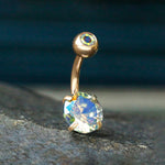 A close-up image of the Kaya Movement Aurora Navel Jewelry, featuring a small round gem on the top ball and a larger, multi-faceted, iridescent aurora borealis crystal at the bottom. This piece, known for its simple yet elegant design, boasts a 316L surgical steel construction with a rose gold finish. The jewelry rests on textured stone with a blurred dark background.