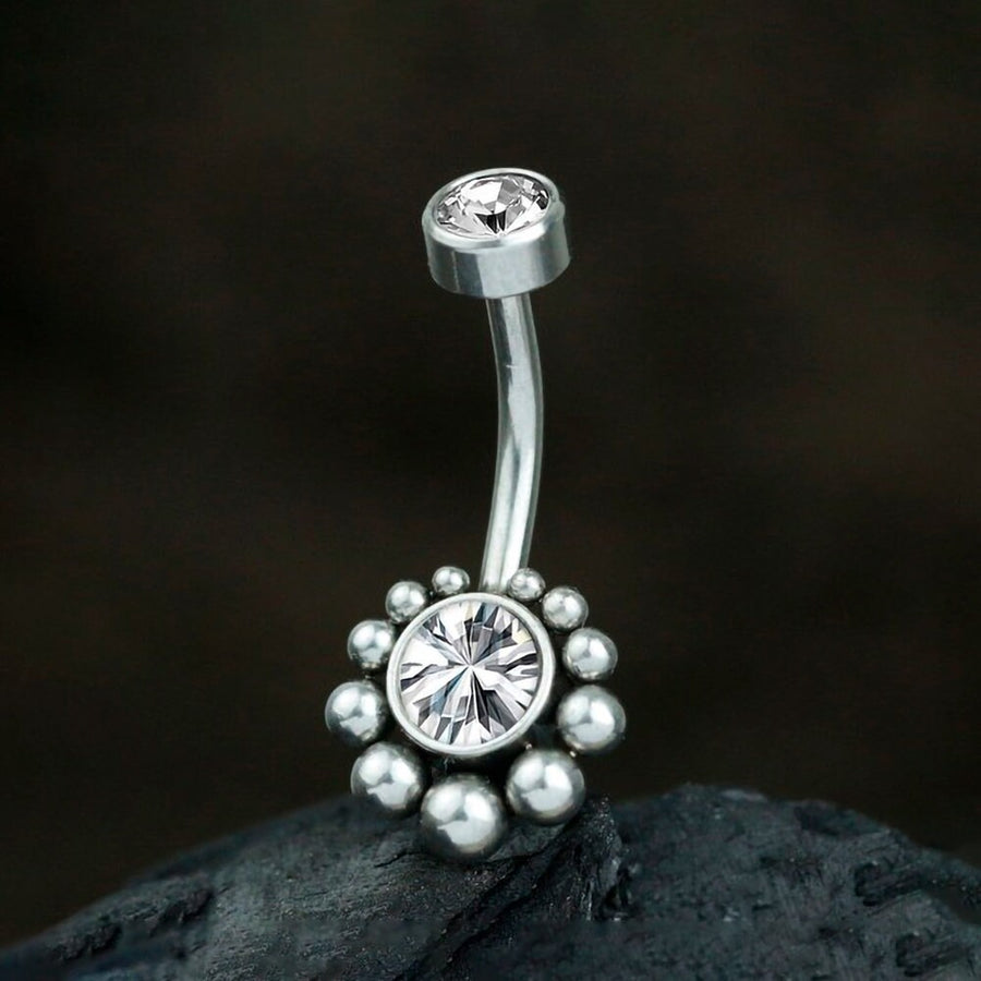 A close-up shot features Kaya Movement's Zirka Navel Jewelry - Boho Essence with Sparkling Zircon, crafted from silver 316L surgical steel. The exquisite belly button ring showcases two clear gemstones; a smaller one sits at the top, and the larger stone is nestled in a decorative flower-like design at the bottom. This elegant piece of natural and bohemian navel jewelry is displayed on a dark, textured surface.