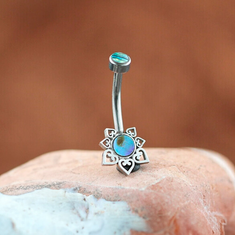 The Mandara Navel Piercing by Kaya Movement is a boho-chic mandala belly jewel crafted from discreet hypoallergenic steel. It features a curved barbell adorned with an abalone shell at one end and heart cut-outs around the iridescent center. The other end sparkles with a blue stone, all beautifully showcased against a smooth, multi-colored rock and blurred brownish background.