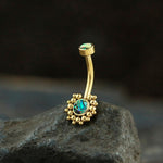 The Solis Navel Piercing by Kaya Movement, exhibiting a geometric golden design and boho shine, is seen resting on a dark rock. The curved barbell's top boasts a small abalone shell at its end, while the bottom displays a larger abalone shell encircled by tiny spherical gold embellishments, all adorned with a luxurious gold finish.