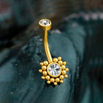 A close-up of Kaya Movement's Lustra Navel Piercing on a dark, textured background. The ring exudes contemporary elegance with its geometric design and two clear, round zircon gemstones; a larger one set in a circular cluster of small gold beads at one end, and a smaller one set in a simple stainless steel gold finish at the other.