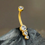 Close-up of the Kaya Movement Xane 1.6mm Navel Piercing adorned with four sparkling zircons, featuring a brilliant gold finish. The jewelry is displayed on a textured gray rock, contrasting beautifully with the smooth and shiny surfaces of the metal and gems.
