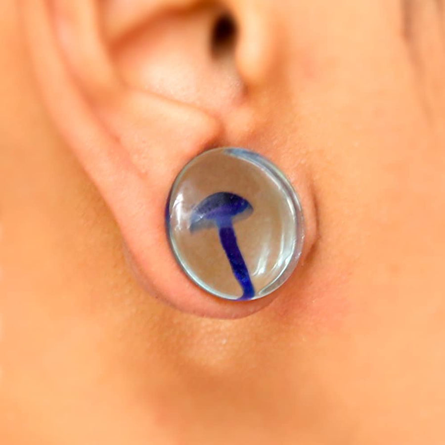 A close-up of a person's ear wearing the Kaya Movement's Psylia Plug - Blue Mushroom Glass Design, featuring a small blue mushroom piece embedded inside. The transparent ear plug is perfectly stretched to fit the earlobe, with a blurred background emphasizing the intricate details of the ear plug. This Psylia Plug is available in sizes ranging from 6mm to 16mm (2g to 5/8"), embodying a true psychedelic festival vibe.