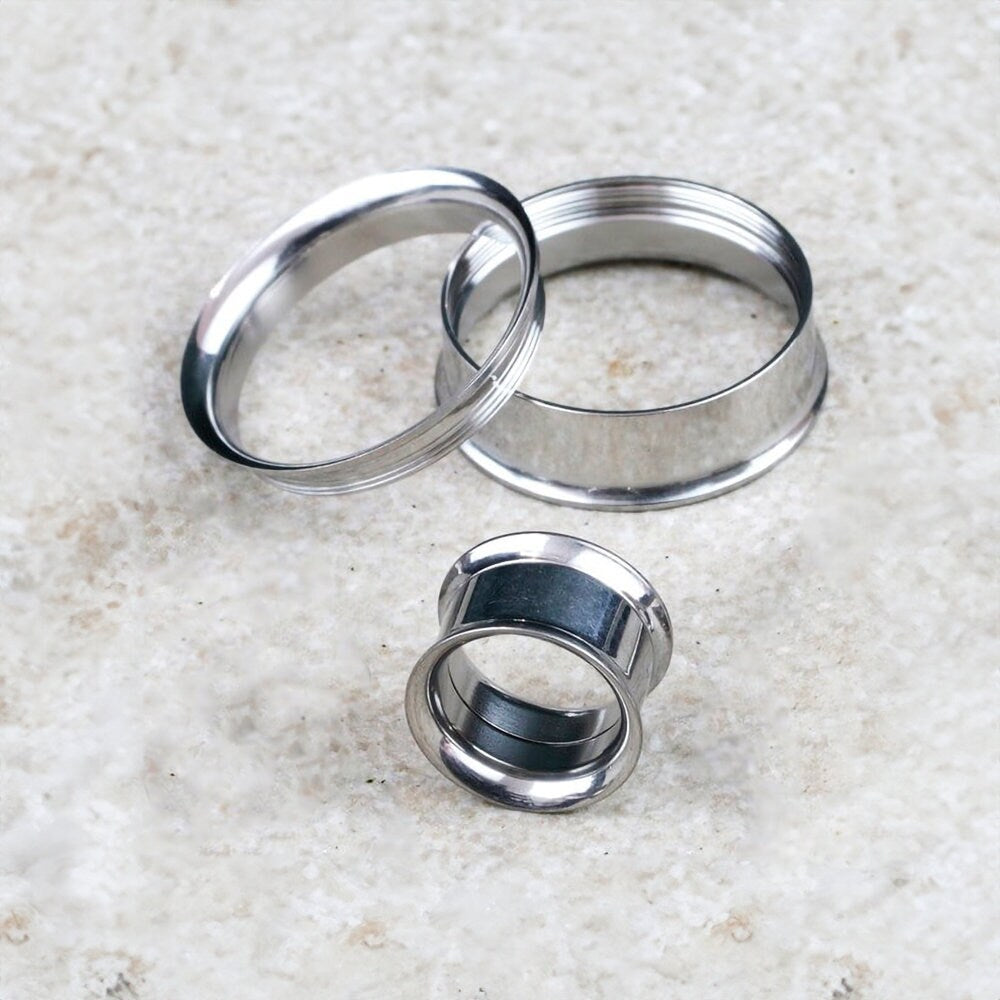 Three Kaya Movement Zura Tunnel ear plugs of varying sizes are scattered on a textured white surface. One ear plug stands upright while the other two lie on their sides, showcasing the minimalist charm and unique angles of these 316L surgical steel jewelry pieces in silver finish.