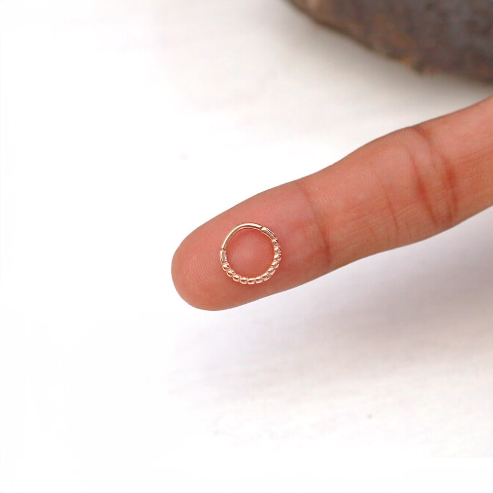 A hand with a finger outstretched holds a small, rose gold Liora Elegant Septum ring by Kaya Movement delicately balanced on the fingertip. The background is white, and the stainless steel 316L ring is detailed with a pattern of small, round beads along part of its circumference.