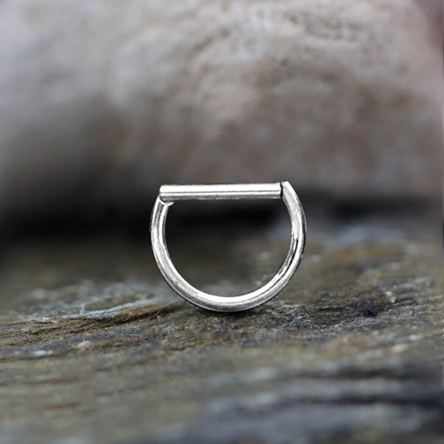 A close-up of the Anneau Segmenté Argenté 10mm Minimalist by Kaya Movement, a sleek silver D-shaped nose ring designed as a piercing clicker, resting on a textured surface. The background is blurred, emphasizing the fine craftsmanship and simple elegance of the jewelry ideal for daily wear.