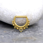 A small gold septum ring from Kaya Movement, known as the Anneau de Septum en Acier Inoxydable 316L with Placage Or PVD, features a 1.2mm bar and a 10mm inner diameter. Adorned with a row of small spherical beads and three heart-shaped charms, it is elegantly displayed against a smooth, rounded white stone. The fine craftsmanship of the PVD gold plating and bead details gives it a delicate and elegant appearance.