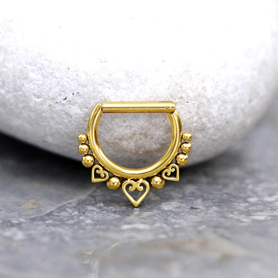 A small gold septum ring from Kaya Movement, known as the Anneau de Septum en Acier Inoxydable 316L with Placage Or PVD, features a 1.2mm bar and a 10mm inner diameter. Adorned with a row of small spherical beads and three heart-shaped charms, it is elegantly displayed against a smooth, rounded white stone. The fine craftsmanship of the PVD gold plating and bead details gives it a delicate and elegant appearance.