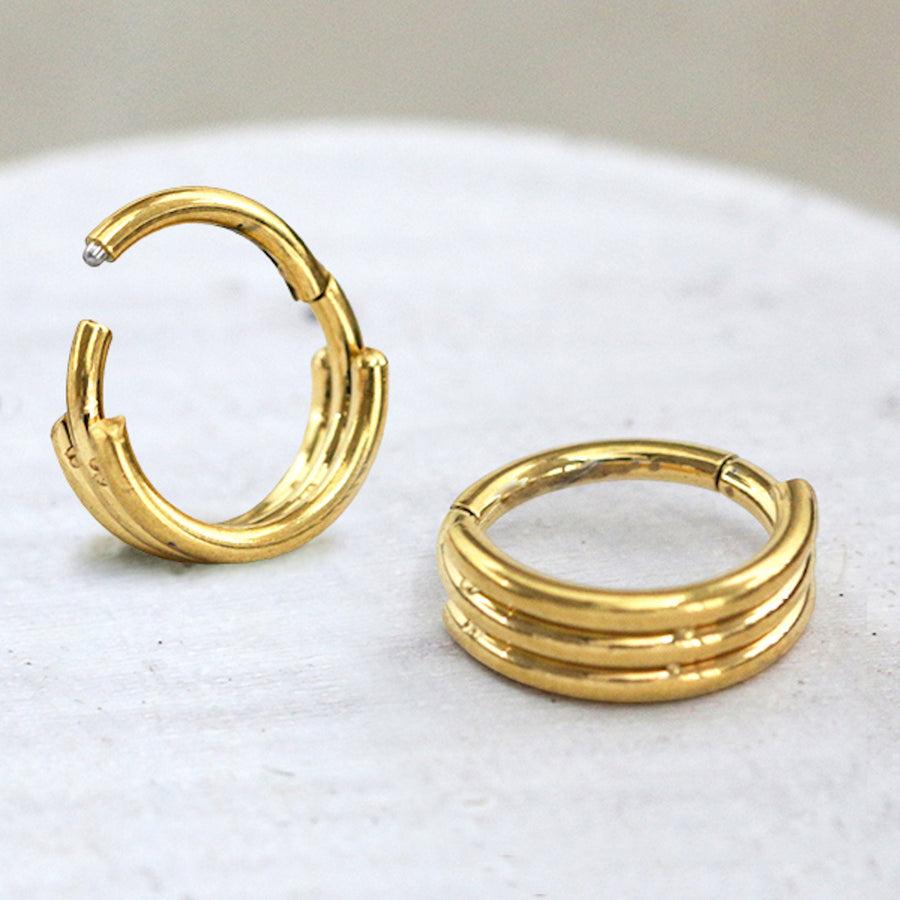 Close-up view of the Anneau 8mm Segmenté en Or pour Piercing from Kaya Movement, featuring a minimalistic three-band design, crafted from acier 316L. One earring stands upright while the other lies flat on a white surface. Their segmenté à charnière details add elegance to this versatile piece suitable for septum, tragus, and helix piercings. A blurred neutral background emphasizes their beauty.