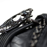 Close-up of a metallic skull-shaped keychain attached to the APOPHIS Post-Apocalyptic Leather Shoulder & Belt Bag from Kaya Movement, adorned with black studs. The focus is on the detailed skull with visible teeth and eye sockets, while the background is slightly blurred, adding an edgy touch perfect for any wasteland festival accessory.