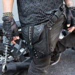 A person wearing the APOPHIS Post-Apocalyptic Leather Shoulder & Belt Bag from Kaya Movement, adorned with black studs, stands beside a motorcycle. They are dressed in black jeans, a patterned black shirt, and fingerless gloves with multiple bracelets on their wrist, exuding the perfect vibe for a wasteland festival accessory.