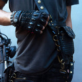 A person wearing a studded leather glove, an arm strap with metal rings and studs, and holding a Kaya Movement SHIPIBO Tribal Apocalyptic Leather Belt & Shoulder Bag | Labradorite adorned with metal details. They are standing next to a motorcycle, partially visible in the background.