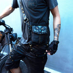 A person wearing a dark t-shirt and black shorts stands next to a motorcycle. They sport the SHIPIBO Tribal Apocalyptic Leather Belt & Shoulder Bag with Labradorite by Kaya Movement, stylishly wrapped around their chest and waist. The person has a tattoo on their right forearm and is wearing a fingerless glove on their left hand, ready for any dystopian festival adventure.