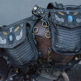 A close-up view of two intricately designed SHIPIBO Tribal Apocalyptic Leather Belt & Shoulder Bags by Kaya Movement, featuring metal clasps and Labradorite gemstone decorations. The bags are strapped onto a piece of weathered, rustic metal equipment with brass rings and fastenings, evoking the rugged charm suited for dystopian festivals. The background is a neutral grey wall.