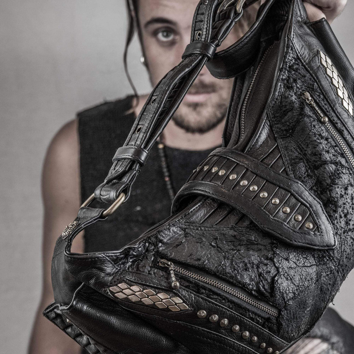 A person with long hair and a beard partially visible in the background holds up the rugged and stylish Nebula Unisex Leather Off Shoulder Cross Body HandBag from Kaya Movement, featuring intricate detailing, studs, and zippers that showcase its design. The plain and neutral backdrop contrasts sharply with the dystopian festival vibe of this accessory.