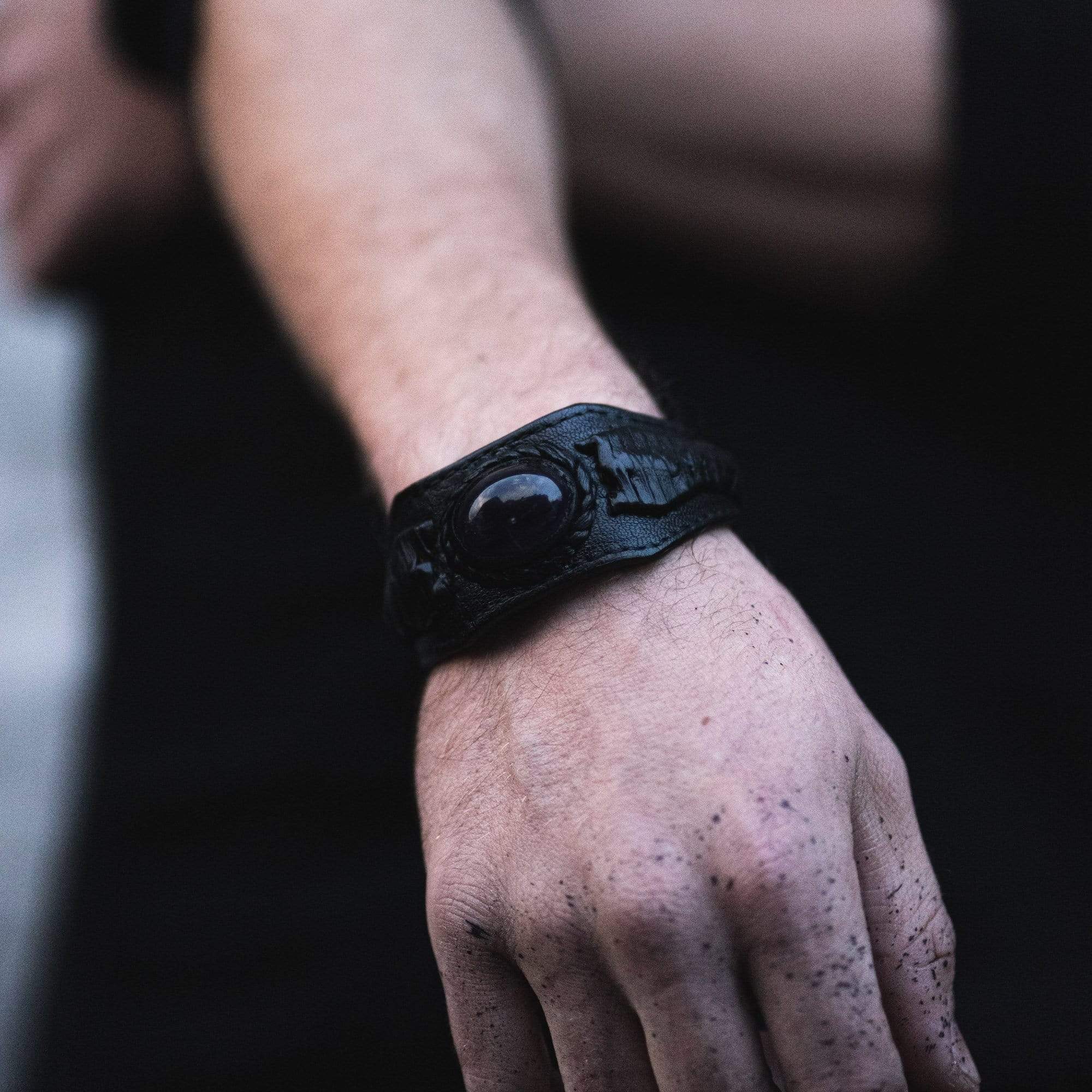 Close-up of a person's left hand and wrist, adorned with the Kaya Movement ANOM Men's Black Cuff Leather Bracelet featuring a central amethyst stone. The person's skin is lightly speckled with what seems to be dark dust or paint, contrasting against a dark, blurred background.