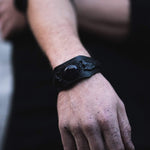 Close-up of a person's left hand and wrist, adorned with the Kaya Movement ANOM Men's Black Cuff Leather Bracelet featuring a central amethyst stone. The person's skin is lightly speckled with what seems to be dark dust or paint, contrasting against a dark, blurred background.
