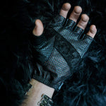 A hand wearing a black, fingerless glove with textured details and a lace pattern is displayed against a dark, furry background. The wrist is adorned with the Kaya Movement's KRAKO Reptilian Spine Cuff Bangle in Gold or Black, crafted using the lost wax process. The glove contrasts with the background, highlighting its intricate design.