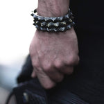 Close-up of a person's hand wearing a spiked bracelet and holding a folded black object. The person is dressed in dark clothing, accentuating the KRAKO Reptilian Spine Cuff Bangle in Gold or Black by Kaya Movement on their wrist. The background is blurred, emphasizing the accessories' details and adjustable size.