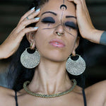 A person with closed eyes wears SAYAGATA Oversized Tribal Hoop Disc Earrings in Gold by Kaya Movement, a choker necklace, and facial piercings. Their face is adorned with bold makeup and symmetrical decorative lines, including a crescent moon symbol on their forehead. Hands touch their head gently.
