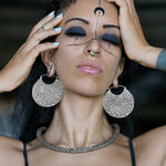 A person with elaborate face paint, piercings, and SAYAGATA Oversized Tribal Disc Hoop Earrings in Silver by Kaya Movement holds their head with both hands. They have closed eyes, dark makeup, long nails, and a woven choker necklace, exuding a look of intense concentration or meditation.