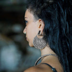 A person with long, dark hair is seen from the side wearing large, intricate SAYAGATA Oversized Tribal Disc Hoop Earrings in Silver by Kaya Movement which feature a maze-like pattern. They also have a nose piercing and are adorned with a textured necklace. The background is blurred and indistinct.