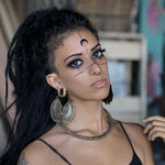 A woman with long dark hair styled in a high ponytail is wearing the CIRCUIT Oversized Visionary Disc Hoop Earrings in Gold by Kaya Movement. She also has a gold chain necklace on. Her face is adorned with intricate black markings around her eyes and forehead as she looks slightly upwards with a neutral expression.