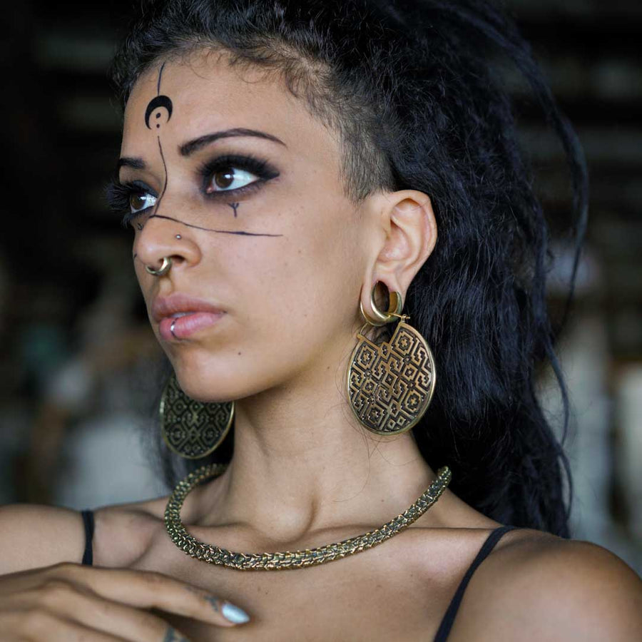 A woman with a contemplative gaze wears large, circular Shipibo-patterned earrings and a matching elegant necklace. Her facial features are accented with a septum piercing and distinct facial markings that complement the geometric designs of her jewelry. Her look is finished with dramatic eye makeup and a naturally poised stance, evoking a strong connection to both tradition and contemporary style.