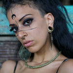 A woman with dark, long hair looks at the camera. She has a side shave haircut, wears large earrings, a Kaya Movement DARMA Biomechanical Clicker Hoop Septum Nose Ring in Gold (16 gauge), and a chain necklace. Her face is adorned with black geometric makeup and a half-moon symbol on her forehead. She is wearing a black top.