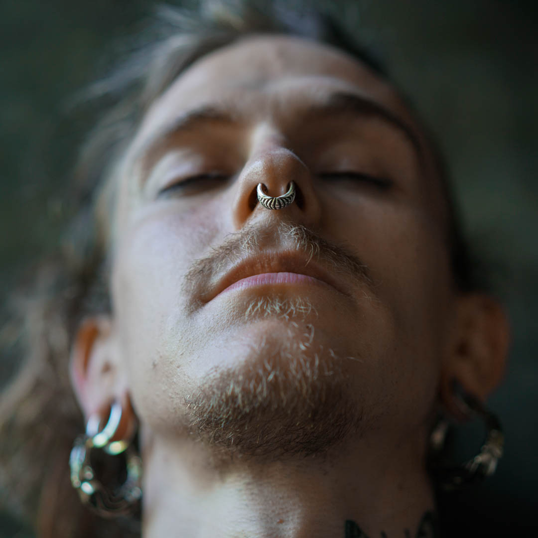A close-up of a person's face looking upward with their eyes closed. They are wearing the Kaya Movement Darma Biomechanical Clicker Hoop Septum Nose Ring in Silver, 16 gauge, and have stretched ear lobes with tunnels. Facial hair includes a mustache and a goatee, giving them a rugged edge. The blurred, dark background adds to the post-apocalyptic vibe.