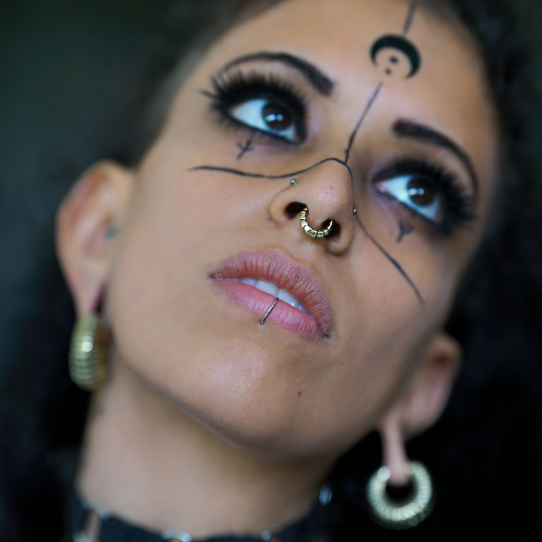A person gazes upward with dramatic facial makeup featuring black lines and symbols. They have multiple piercings, including an AARO Cyborg Seamless Hoop Septum Nose Ring in Gold by Kaya Movement, a lower lip piercing, and ear gauges. They are also wearing large, intricate biomechanical earrings and have defined eyelashes.