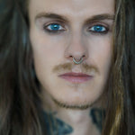 A person with long brown hair and striking blue eyes, wearing the Kaya Movement's AARO Cyborg Clicker Hoop Septum Nose Ring in Silver and subtle makeup, stares directly into the camera with a neutral expression. They have light facial hair and tattoos are faintly visible on their neck, giving off a hint of dystopian body jewelry.
